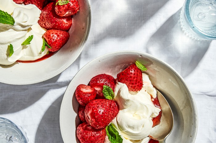 Two white bowls filled with strawberries, cream and green herbs on a white table cloth