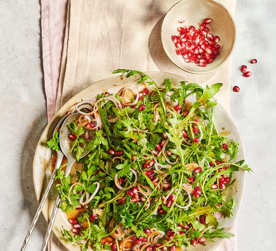 A round white serving plate of green leaves and red pomegranate seeds, sat on a folded piece of white linen