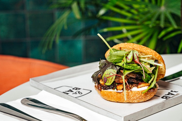 a bread roll filled with aubergine slices sat on a tray with plants in the background