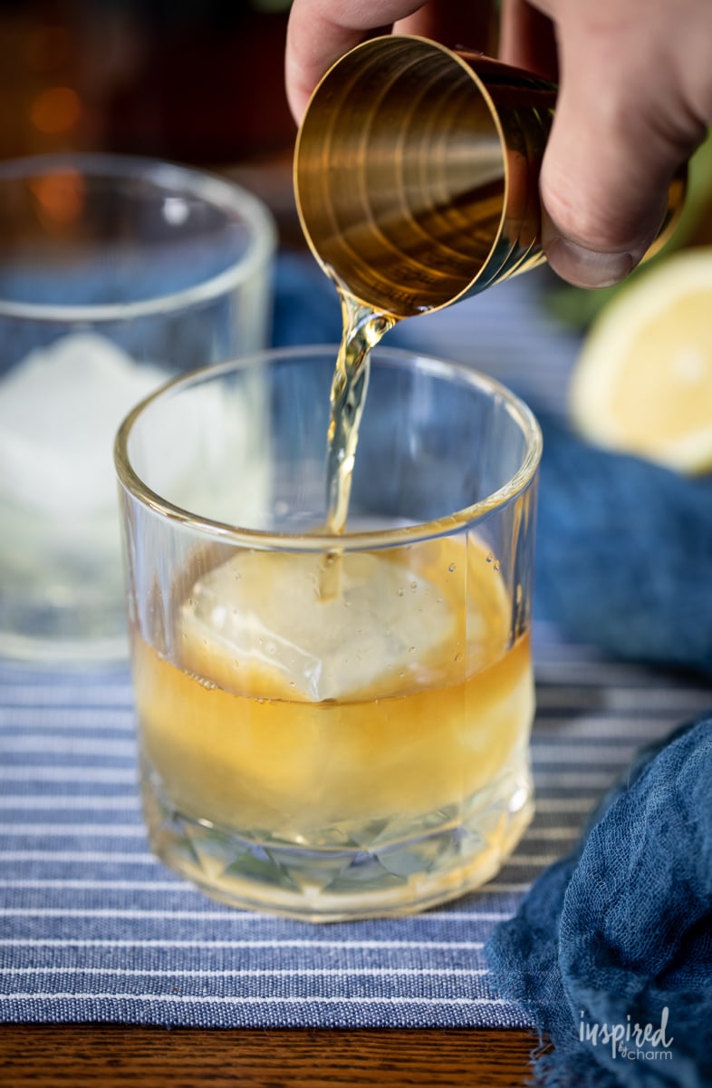 hand adding bourbon to a rocks glass with an ice cube. 