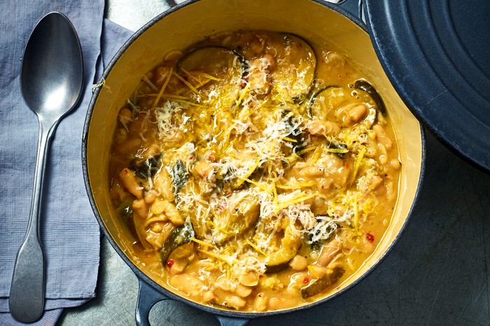 A cast iron pot of creamy braised courgette with the lid leaning on the side, on a grey concrete background with a grey napkin and brushed sliver serving spoon