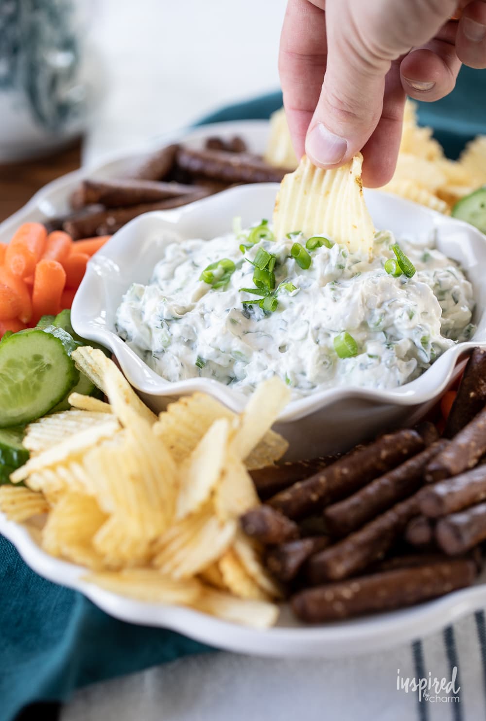 hand with chip in green onion dip in a bowl on a plate with chips and vegetables.
