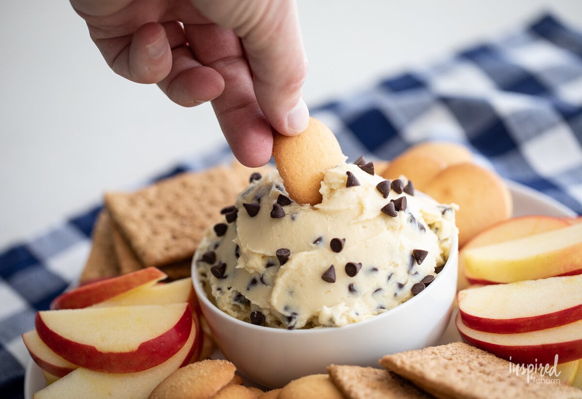 hand dipping Nilla wafer into chocolate chip cheesecake dip in a bowl with cookies and apple slices.