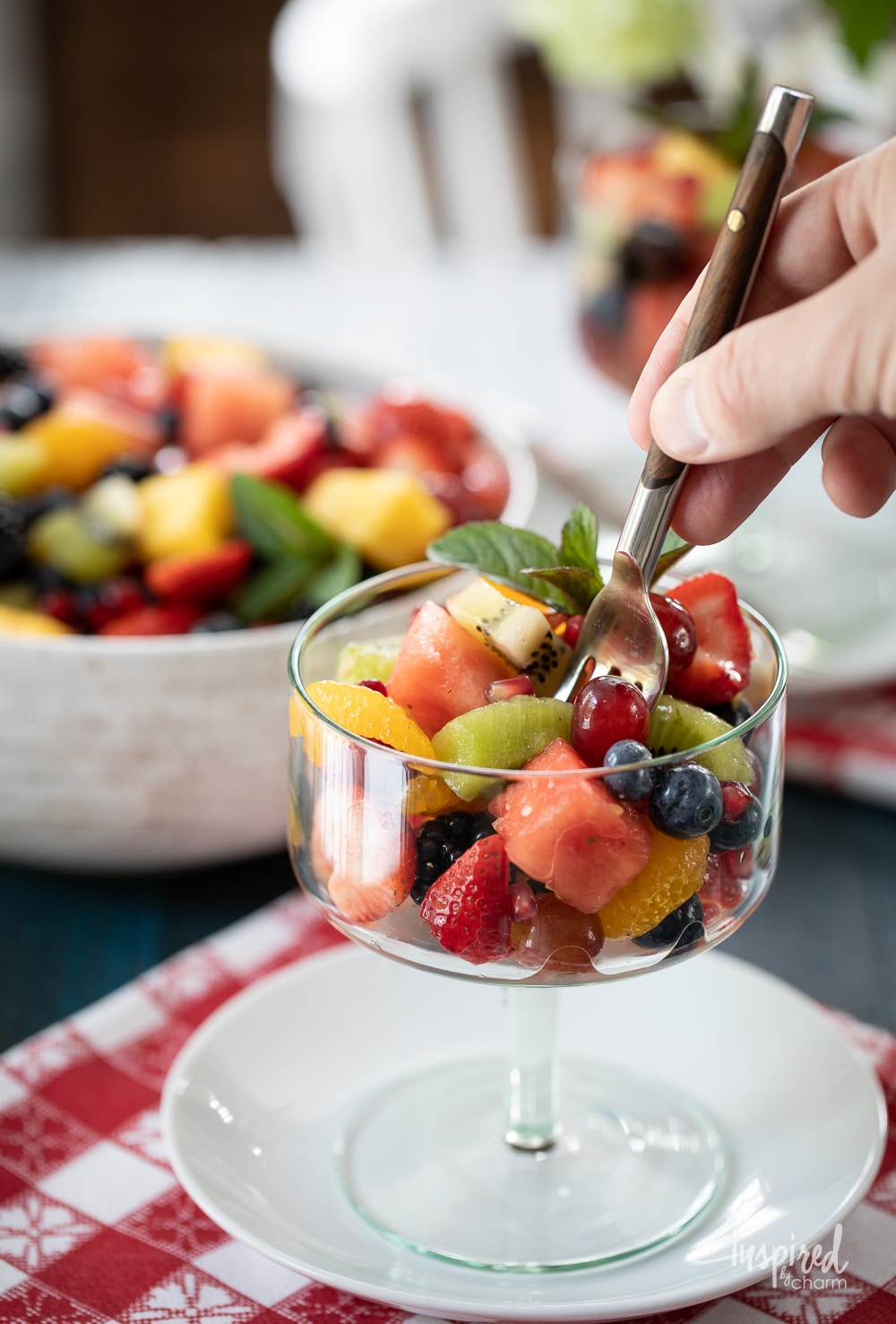 hand taking a bite of fruit salad with a fork from a glass dish.