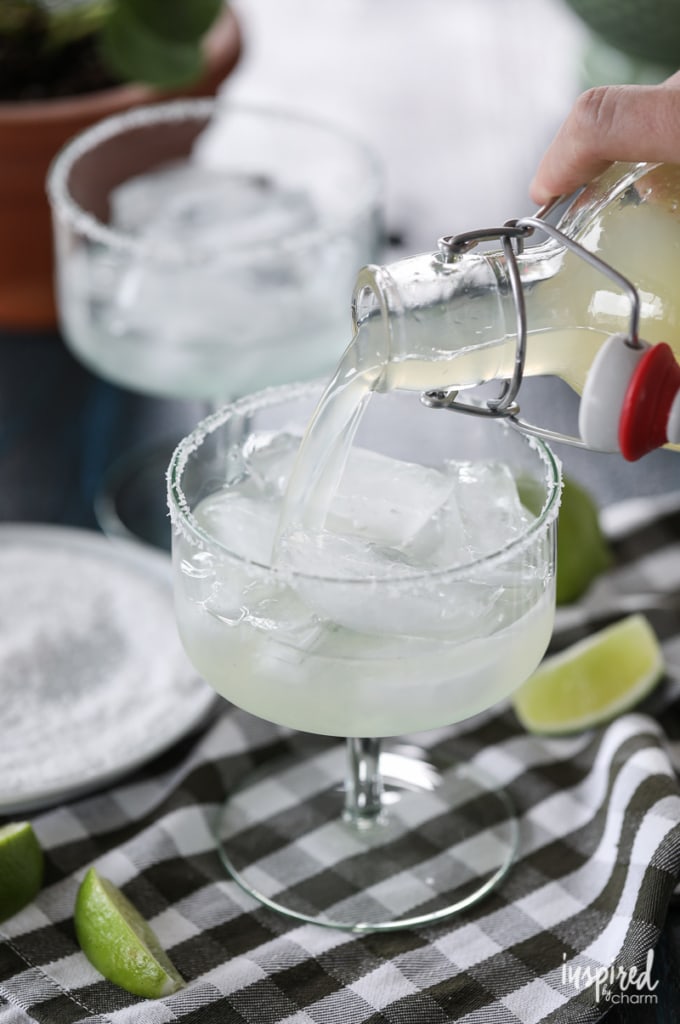 hand pouring a bottle of omemade margarita mix into a cocktail glass with ice.