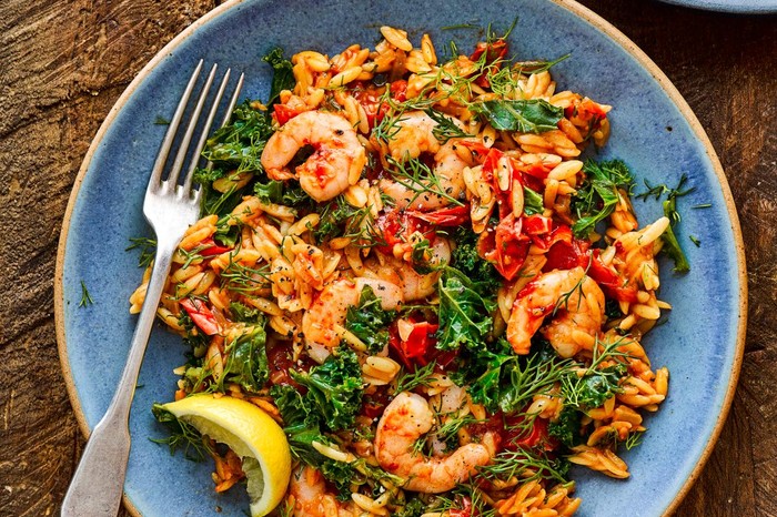 Baked orzo with harissa prawns on a blue plate with a wedge of lemon and fork