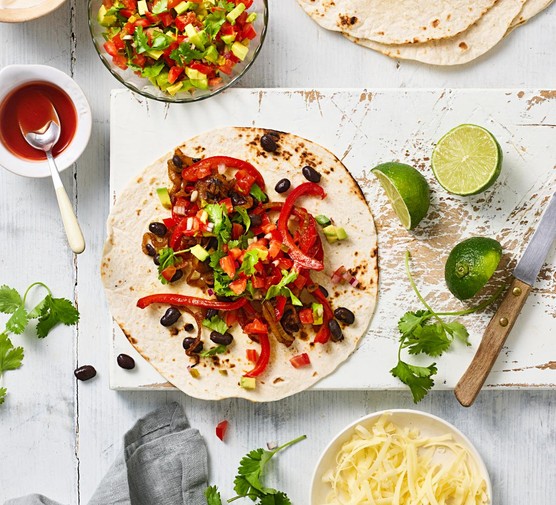 Veggie fajitas with beans, peppers, avocado, coriander and lime, on a chopping board with lime wedges and a knife